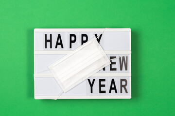 Lightbox with text HAPPY NEW YEAR with medical mask on green background. New year celebration during coronavirus pandemic. Top view