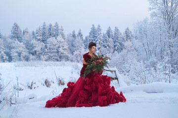 Fototapeta na wymiar A young girl in a lush Marsala dress with a large bouquet of red anemones on the background of a snow-covered landscape