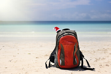 A red sports backpack lies on the ocean shore on snow-white sand with a Santa Claus hat on top. The traveler's backpack is on the sand. Santa's hat is attached to the bag
