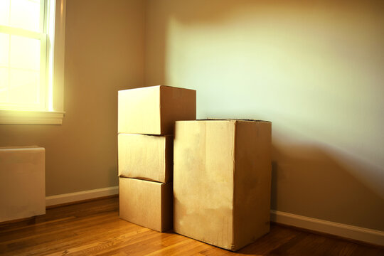 Cardboard moving boxes in the corner of an empty room of a new house. Light shines through a nearby window onto hardwood floors and blank walls.