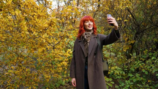 a girl with red hair in a coat, takes a selfie against the background of autumn foliage in the park and then posts it to the network