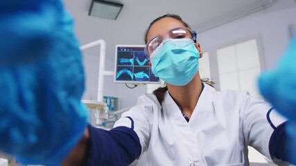 Patient point of view to dentist in protective mask holding tools examining person with toothache sitting on stomatological chair while nurse preparing tools for surgery.