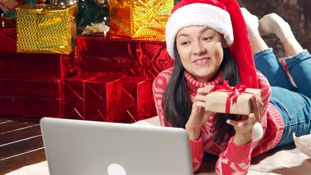 Young happy woman in Santa hat giving gift and talking on communication video using laptop lying on floor at home. Social distancing, new normal and holiday concept