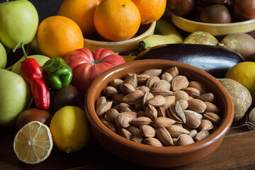 A homemade clay bowl filled with whole almonds in their shell on an old wooden table with fruits, vegetables, and other organic and natural ingredients in the background. Nature and healthy food.