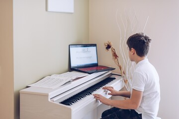 12 year old boy playing the white piano looking at the notes of his laptop.