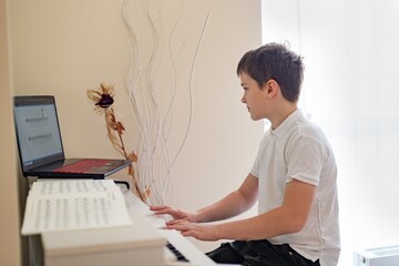 A young boy is learning to play the white digital piano, he looks at the sheet music in his laptop.