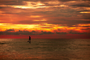 Fototapeta premium Sea sup surfing under amazing dark sunset sky. Two people on Stand Up Paddle Board. Orange sky. Paddleboarding Concept. Phuket. Thailand.