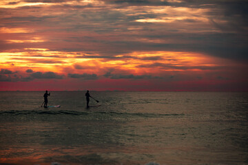 Fototapeta premium Sea sup surfing under amazing dark sunset sky. Two people on Stand Up Paddle Board. Orange sky. Paddleboarding Concept. Phuket. Thailand.