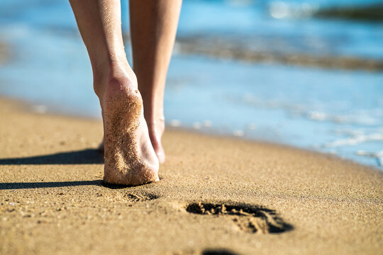 Close Up Of Woman Feet Walking Barefoot On Sand Leaving Footprints On Golden Beach. Vacation, Travel And Freedom Concept. People Relaxing In Summer.