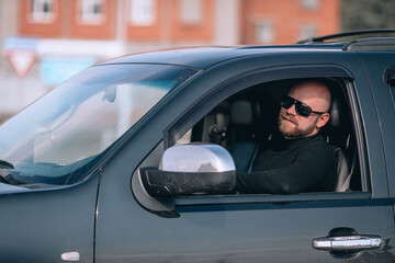 bald and bearded man in glasses with a clock in a suit behind the wheel of a black car
