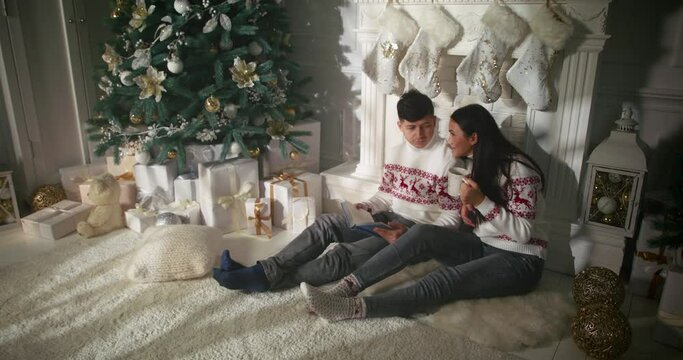 Young Caucasian couple relaxing near fireplace, resting together on sunny warm Christmas morning. Husband reading a book, wife drinking tea, while sitting on floor near xmas tree.