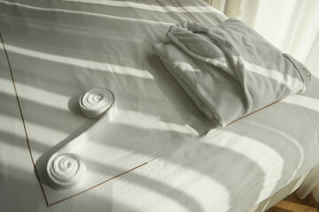 Folded white bathrobe on the bed in hotel room. Clean folded bathrobe on bed in room. Sun shining through the window.