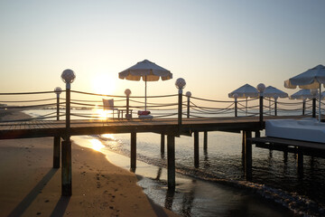 Obraz premium Wooden pier with sunbeds and umbrellas at sunset. Pier with chaise loungers in the sea at resort during sundown. Summer vacation at a sea coastline.