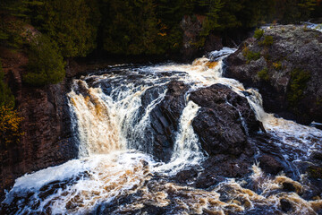 Obraz premium A close up waterfall view of the upper Potato River Falls as the water cascades down and through the rocky riverbed and cliffs surrounded by evergreen trees in the wilderness.