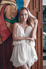 Portrait of smiling blonde girl with evening make up in a white stylish short dress. Beautiful model woman standing near colorful wall. Phuket. Thailand.