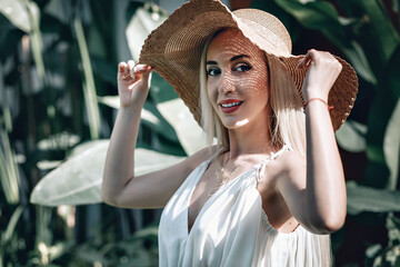  portrait of Young blonde girl with bright make-up in white dress and straw hat at the street posing at the camera and enjoying the sun. Close up photo.