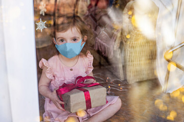 Little beautiful girl in a medical mask is sad at the window of the house, holding a New Year's gift in her hands. The baby won't get to make a wish to Santa Claus. Christmas Holidays in Quarantine.