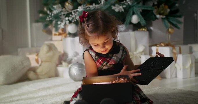 Adorable little girl preschooler in plaid dress opening Christmas gift from Santa Claus, enchanted by bright shiny present toy inside. Xmas tree on background.