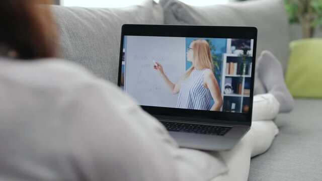 Back view of woman having online course on laptop while lying on grey couch. Female in domestic outfit studying remotely while staying at home.