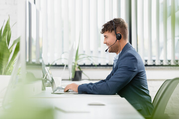  man in headset typing on laptop, while sitting at workplace with plants in office on blurred...