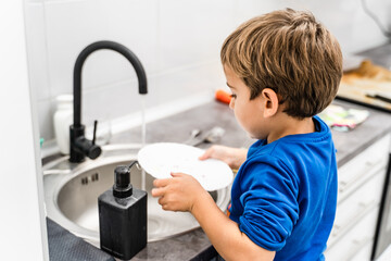 Side view of unknown caucasian boy or girl standing by the sink in the kitchen washing dishes - kid playing while helping in housework -growing up independence concept copy space