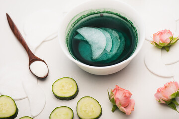 Bowl with cleansing lotion and cotton pads near sliced cucumber, tea roses, and wooden spoon with cosmetic cream on white