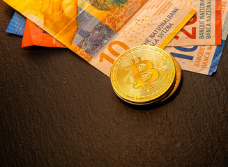 Shiny gold, silver and copper Bitcoin coins lying on European banknotes - Swiss francs on a black flat slate stone.