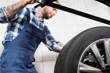 Fototapeta premium tattooed mechanic examining wheel on balance control machine in workshop on blurred 