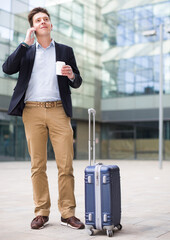 cheerful worker in suit standing with baggage and talking phone