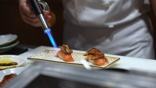Close up of Professional chef hand use butane torch burner burning salmon sushi with foie gras on serving plate. Chef preparing healthy sushi menu on open kitchen counter bar in japanese restaurant.