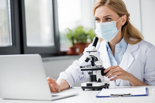 specialist in medical mask using laptop near microscope on desk, stock image
