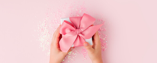 Kids hands holding present with pink bow on pastel pink background. Festive backdrop for holidays: Birthday, Valentines day, Mother day, Christmas, New Year. Flat lay style, sale shopping