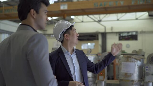 30s young Asian businessman in formal suit and hard hat showing foreign business partner in factory background. Business partnership and team work success concept - 4K Real Time Footage