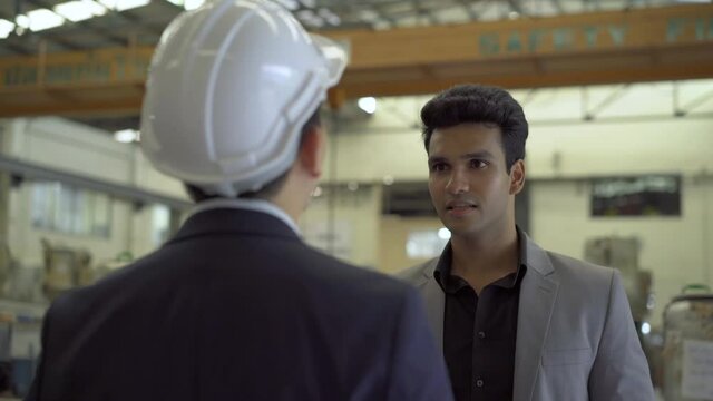 30s young Indian businessman in formal suit and hard hat having business talking in factory background. Business partnership and team work success concept - 4K Real Time Footage
