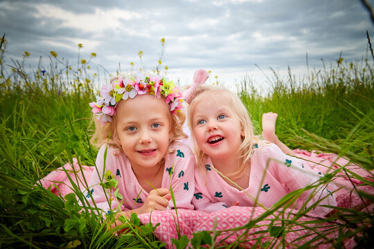 Two pretty young small blonde girls with curly hair in pink dress in field with grass and flowers in summer day. Sisters in photoshoot and cloudy sky background