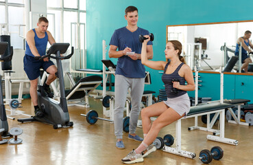 Fototapeta na wymiar Young woman doing exercises with dumbbells with help of instructor at gym