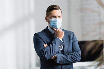  businessman in medical mask looking away, while standing in office on blurred foreground