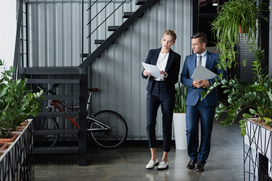 Businessman with laptop looking at paper sheets in hands of female colleague, while standing at work, stock image