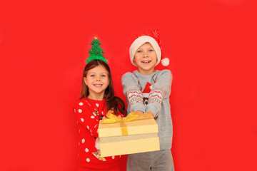 Two children in sweaters are holding a New Year's gift on a red background. A boy and a girl give a Christmas gift to each other