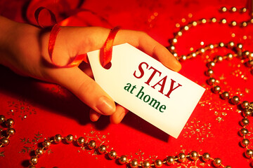 A woman's hand holds a white label with the text - stay at home, on a decorated Christmas background, during the Christmas and New Year holidays. 