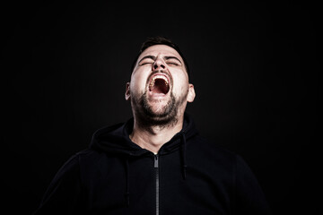 Young man emotionally shouts with his head up. Black background. Stress, pain, aggression. Close-up.