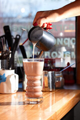 Barista hand pouring milk into a glass with cocoa. Refreshing summer drink
