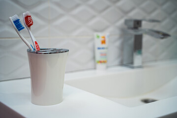 A pair of toothbrushes in a white plastic cup against a background of white tiles in the bathroom. The toothbrushes sit on a white stone sink.