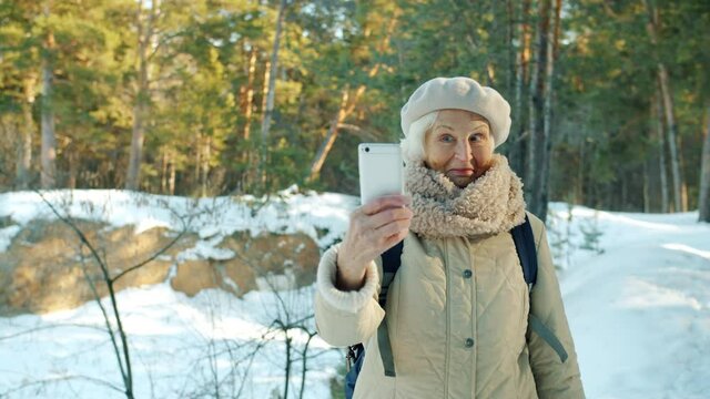 Beautiful senior woman hiker is taking pictures with smartphone camera in winter park posing smiling looking at screen. People and modern devices concept.