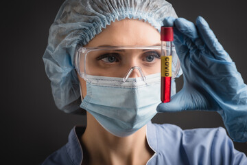 nurse in medical cap, mask and goggles holding test tube with covid lettering on blurred isolated...