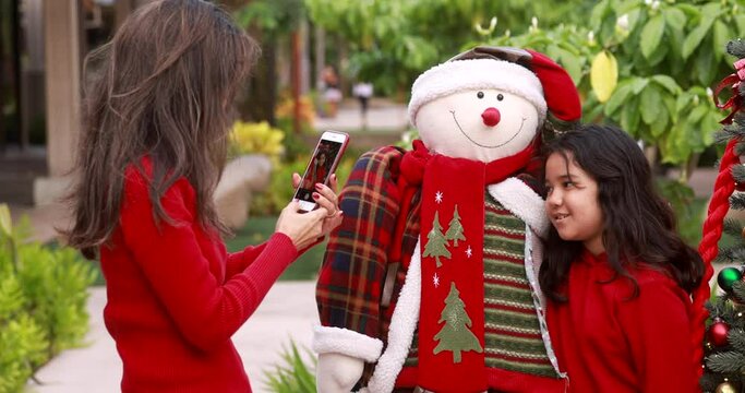 Beautiful exotic asian older woman taking photo of young girl next to stuffed mall Christmas snowman and tree with smartphone. Mature woman is having fun with both; bokeh background.