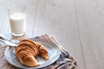 horizontal view of a traditional pastry croissant on a plate with a white and blue napkin and on a wooden table with a glass of milk with a drinking straw on the background