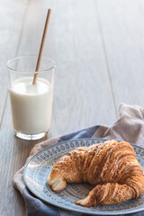 vertical view of a traditional pastry croissant on a plate with a white and blue napkin and on a wooden table with a glass of milk with a drinking straw on the background. copy space on the top.