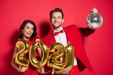  young couple holding disco ball and balloons in shape of 2021 numbers on red background
