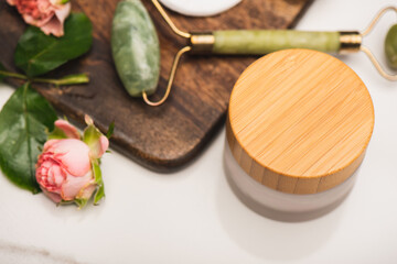 Container of cosmetic cream near chopping board with jade roller, and tea roses on blurred white